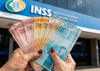 Mãos de pessoa idosa seguram cédulas de dez, vinte e cem reais em frente a uma agência do INSS.