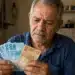 Aposentado segurando uma nota de 100 e outra de 50 reais, com expressão preocupada, representando os pagamentos menores do INSS em junho.