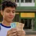 Estudante segura notas de dinheiro em frente à escola, representando os pagamentos de até R$ 1.000 do Pé-de-Meia em maio de 2026.