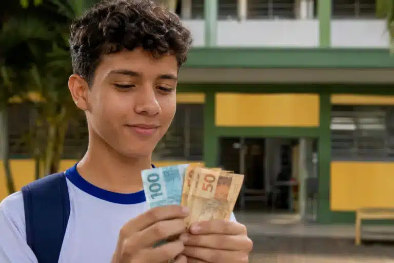 Estudante segura notas de dinheiro em frente à escola, representando os pagamentos de até R$ 1.000 do Pé-de-Meia em maio de 2026.