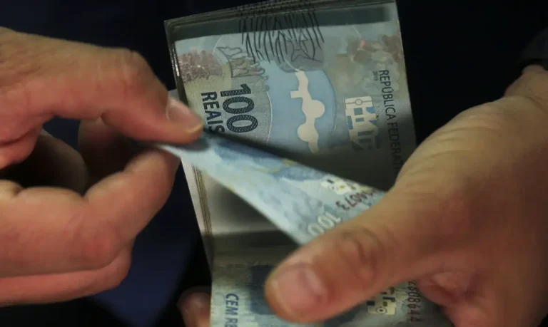 Mãos contam cédulas de cem reais sobre fundo escuro.