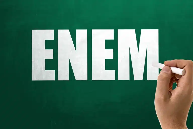 Mão com giz em frente a quadro verde com a palavra Enem em destaque, representando o período de recursos para isenção do Enem 2026.