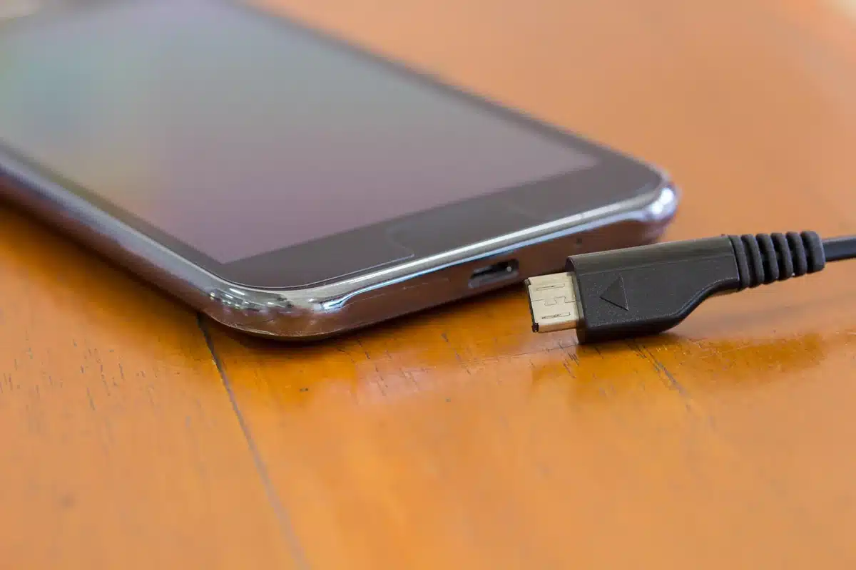 Cabo carregador próximo ao smartphone sobre mesa de madeira, representando a regra dos 20% a 80% para preservar a bateria.