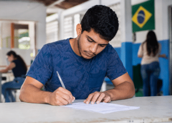 Jovem de camiseta azul escrevendo em folha de prova com bandeira do Brasil ao fundo em ambiente escolar.