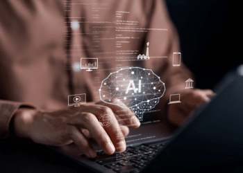 Mãos digitando em laptop com holograma de cérebro AI e ícones de tecnologia flutuando sobre teclado.