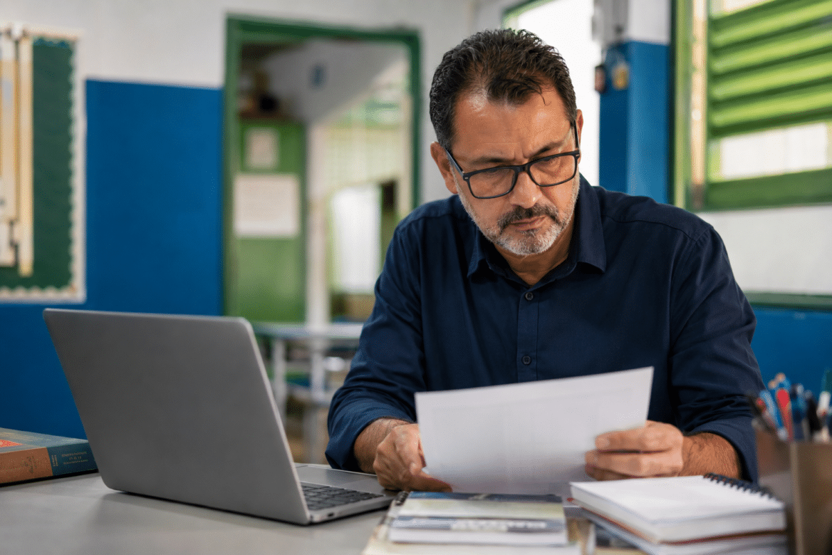 Homem de óculos e camisa azul-marinho lê documento em mesa com laptop, livros e cadernos em sala de aula.