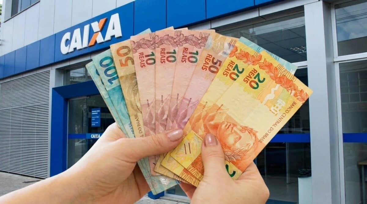 Mãos seguram em leque cédulas de dez, vinte, cinquenta e cem reais em frente a uma agência da Caixa.