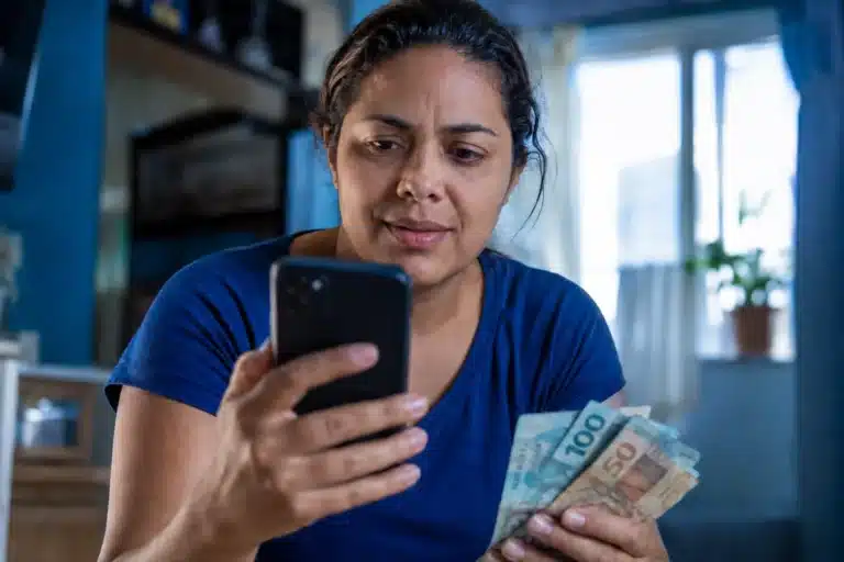 Mulher consulta no celular informações sobre o saque de R$ 6.220 da Caixa, representando a solicitação do valor, que pode ser feita até 30 de maio.
