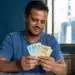 Homem segurando cédulas em ambiente doméstico, ilustrando o saque do FGTS de R$ 6.220 liberado para solicitação a partir de 27 de maio.