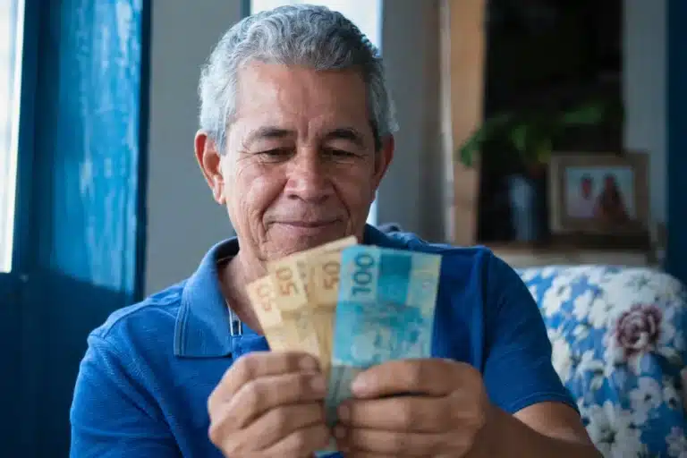 Aposentado brasileiro segurando notas de reais em casa, representando o pagamento da segunda parcela do 13º do INSS.