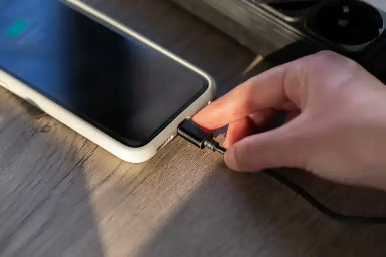 Mão conectando carregador em smartphone sobre mesa de madeira, representando um hábito comum de recarga que pode afetar a vida útil da bateria.