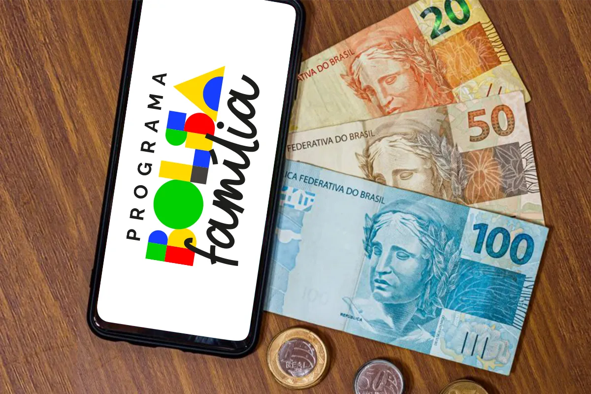 Celular sobre mesa de madeira exibindo aplicativo do programa Bolsa Família ao lado de cédulas de R$ 100 e mão de homem apoiada na mesa