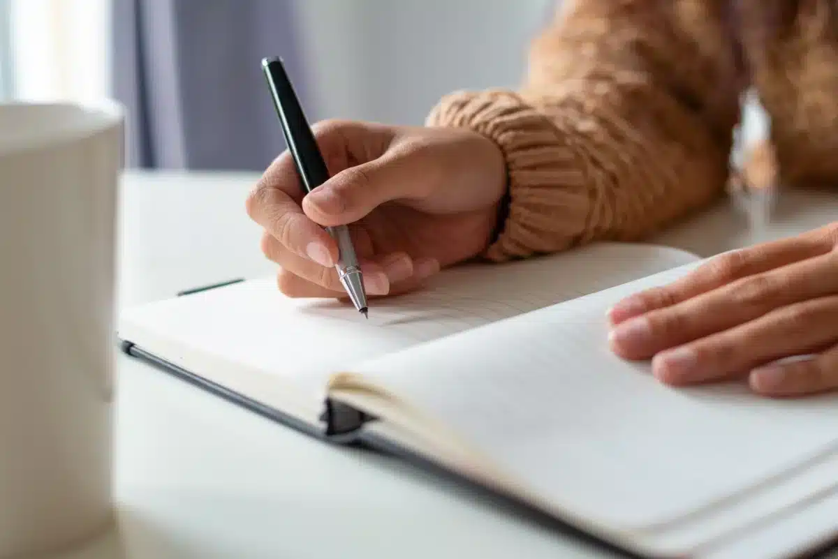 Mão segurando caneta sobre caderno aberto em mesa branca, representando o estudo das regras de pontuação da língua portuguesa.
