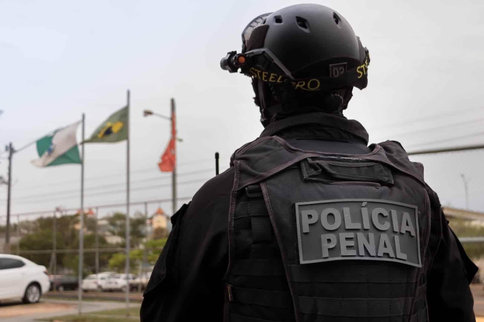 Policial Penal de costas com uniforme tático preto, capacete e óculos de visão noturna, frente a bandeiras do Acre e Brasil