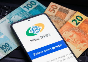 Smartphone exibindo tela do Meu INSS com botão gov.br sobre notas de 100, 50 e 20 reais em teclado preto.