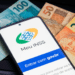 Smartphone exibindo tela do Meu INSS com botão gov.br sobre notas de 100, 50 e 20 reais em teclado preto.