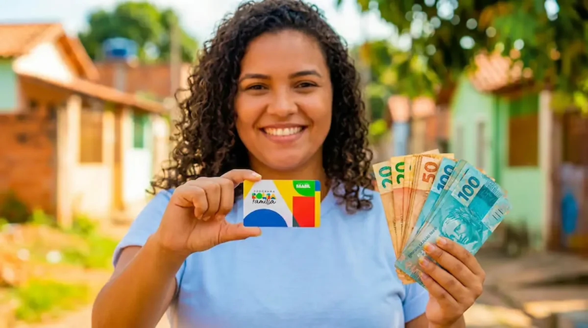 Mulher segurando cartão do Bolsa Família e notas de dinheiro, representando pagamento de junho.