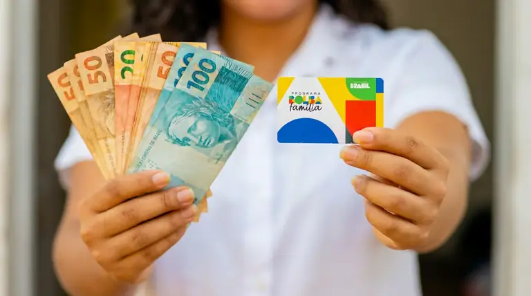 Mãos segurando cartão do Bolsa Família e cédulas de dinheiro, ilustrando pagamento de junho de 2026.
