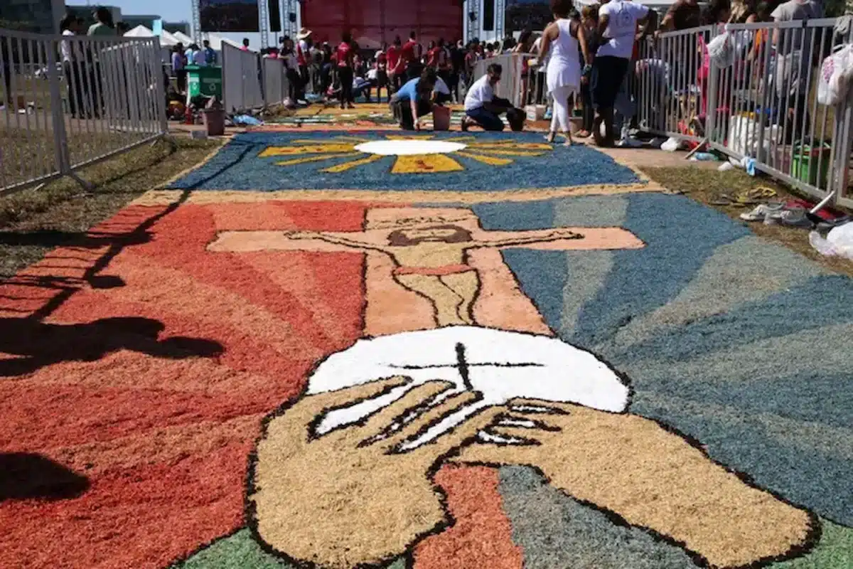 Tapete colorido de Corpus Christi com símbolos religiosos sendo confeccionado por participantes em procissão.