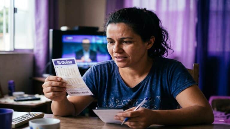 Mulher de camiseta azul segura volante da Quina com caneta na mão, em sala com TV ligada ao fundo