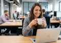 Mulher de blazer cinza segura caneca branca sorrindo diante de MacBook em escritório compartilhado moderno.