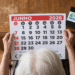 Pessoa idosa com cabelos brancos segura calendário de junho 2026 sobre mesa de madeira, com celular exibindo logo do INSS.