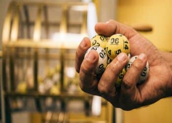 Mão masculina segurando bolas numeradas amarelas e brancas com números 02, 06, 14 e outros, globo ao fundo