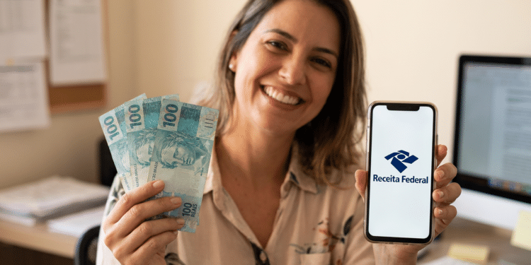 Mulher sorridente segura notas de R$100 e celular com logo da Receita Federal em escritório.