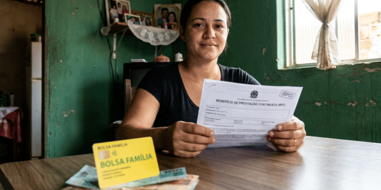 Mulher sentada à mesa segura formulário do BPC ao lado do cartão do Bolsa Família e cédulas de dinheiro