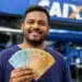 Homem brasileiro sorridente segurando cédulas de reais em frente à agência da Caixa Econômica Federal, ilustrando o saque de R$ 6.220 da Caixa.