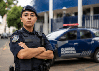Mulher da Guarda Municipal de braços cruzados, farda azul-marinho e colete tático, viatura azul ao fundo.