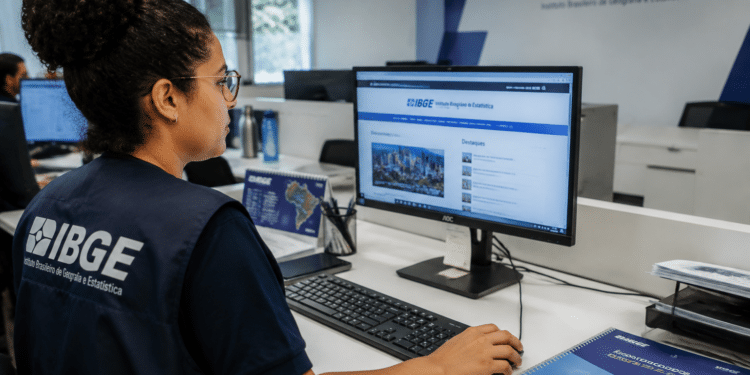 Funcionária do IBGE com colete azul-marinho usa computador acessando site do instituto em escritório com logo na parede.