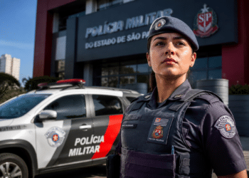 Policial feminina da PMESP usando boina e colete tático posa em frente à viatura e prédio da corporação.