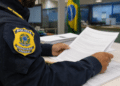 Policial Rodoviário Federal uniformizado lê folhas de documentos em mesa de escritório com bandeira brasileira ao fundo.