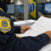 Policial Rodoviário Federal uniformizado lê folhas de documentos em mesa de escritório com bandeira brasileira ao fundo.