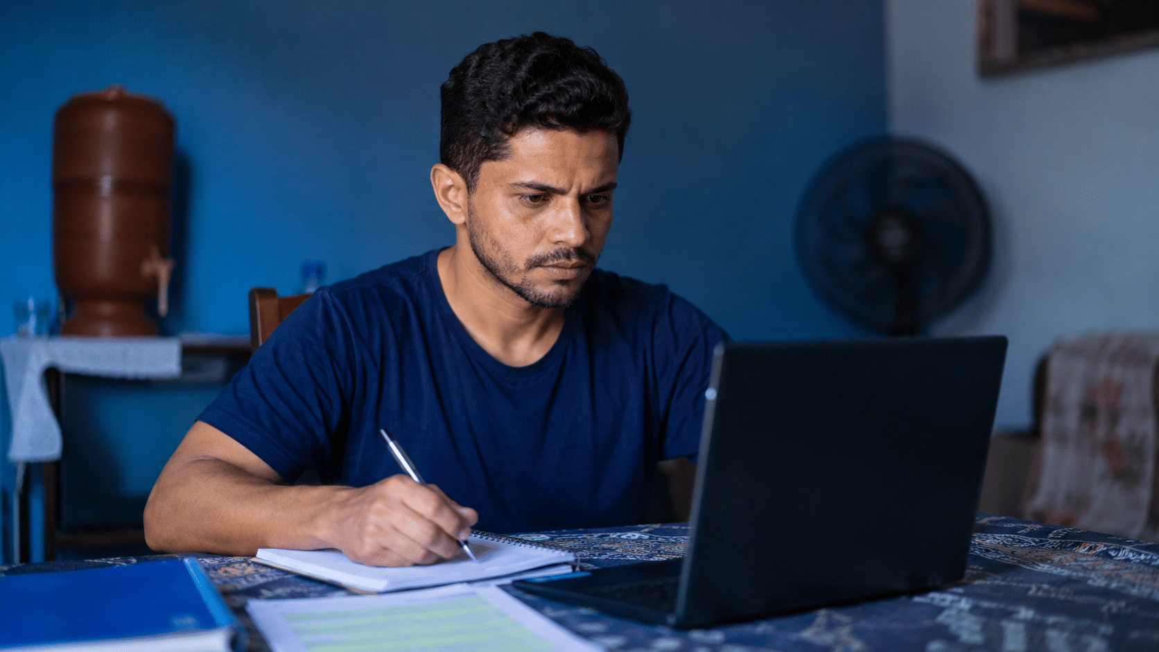 Homem escrevendo em caderno enquanto usa notebook em ambiente doméstico azul.