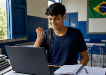 Jovem de camiseta azul marinho celebra com punho cerrado olhando laptop em sala de aula com bandeira do Brasil.