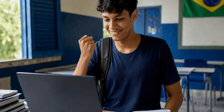 Jovem de camiseta azul marinho celebra com punho cerrado olhando laptop em sala de aula com bandeira do Brasil.