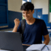 Jovem de camiseta azul marinho celebra com punho cerrado olhando laptop em sala de aula com bandeira do Brasil.