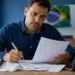 Homem brasileiro revisando documentos do Imposto de Renda 2026 enquanto utiliza o computador, representando a correção de dados enviados à Receita Federal.