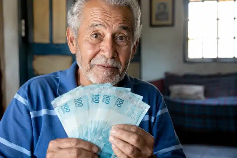 Aposentado brasileiro segurando notas de cem reais em casa, representando os depósitos de R$ 8.475,55 do INSS nesta terça-feira, 2 de junho.