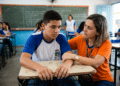 Estudante uniformizado recebe apoio de profissional em sala de aula durante atividade escolar