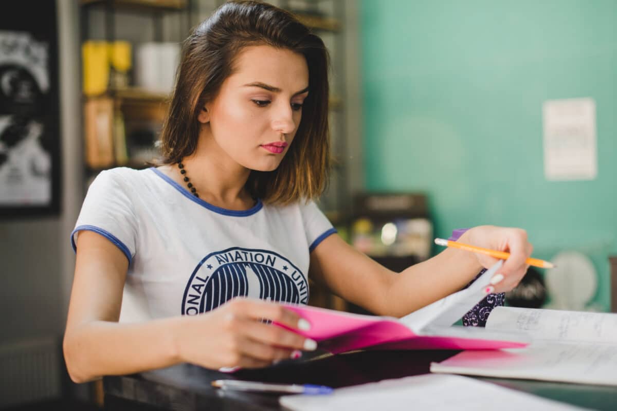 Jovem estudante folheia caderno de estudos sobre mesa em ambiente de biblioteca com livros ao fundo.