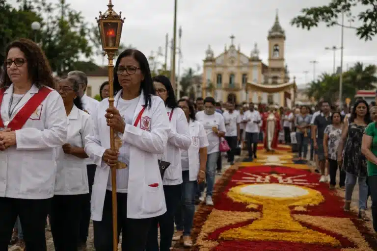 iéis participando de uma procissão de Corpus Christi em rua brasileira, com tapetes coloridos de serragem e flores, representando o feriado de Corpus Christi.