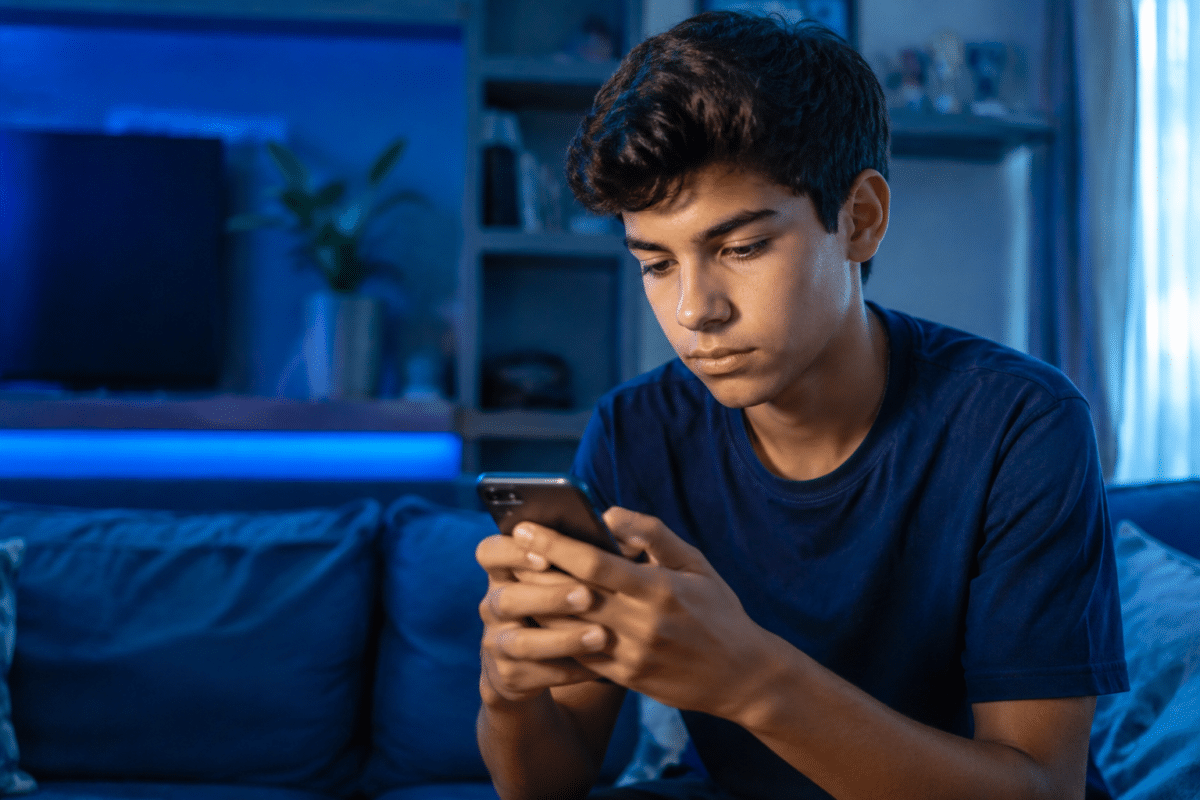 Jovem de cabelos escuros e camiseta azul marinho usa celular em ambiente noturno com iluminação LED azul.