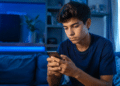 Jovem de cabelos escuros e camiseta azul marinho usa celular em ambiente noturno com iluminação LED azul.