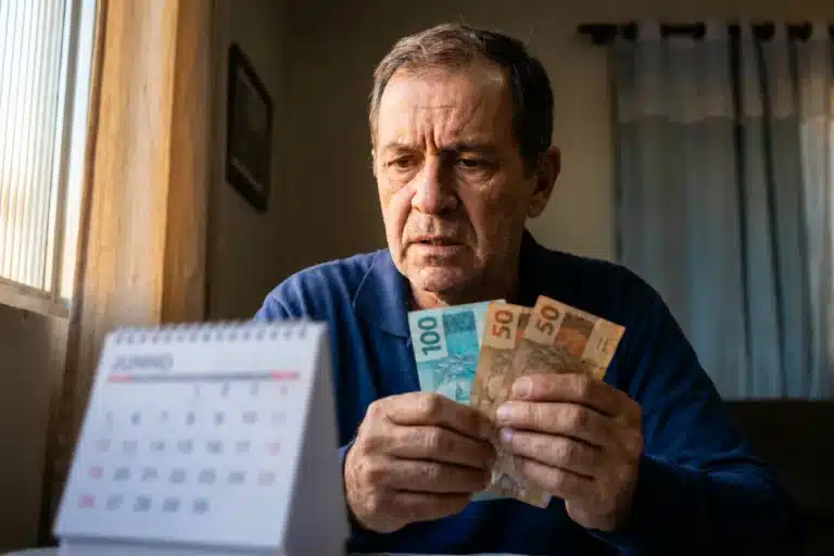 Homem segurando cédulas, olhando para o calendário, representando o fim do adicional de 50% no benefício do INSS em junho.