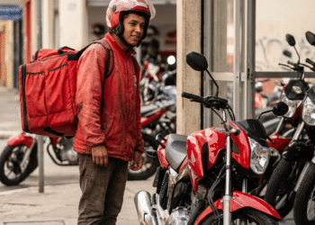 Entregador com mochila térmica observa moto vermelha, veículo que poderá ser financiado pela nova linha de crédito do governo
