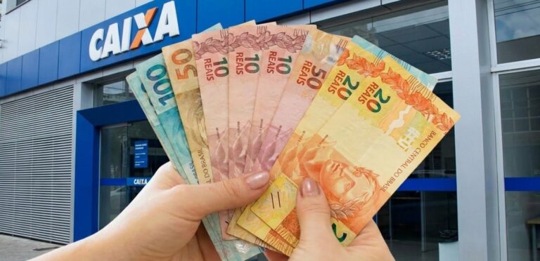 Mãos seguram cédulas de real variadas em frente à agência da Caixa Econômica Federal