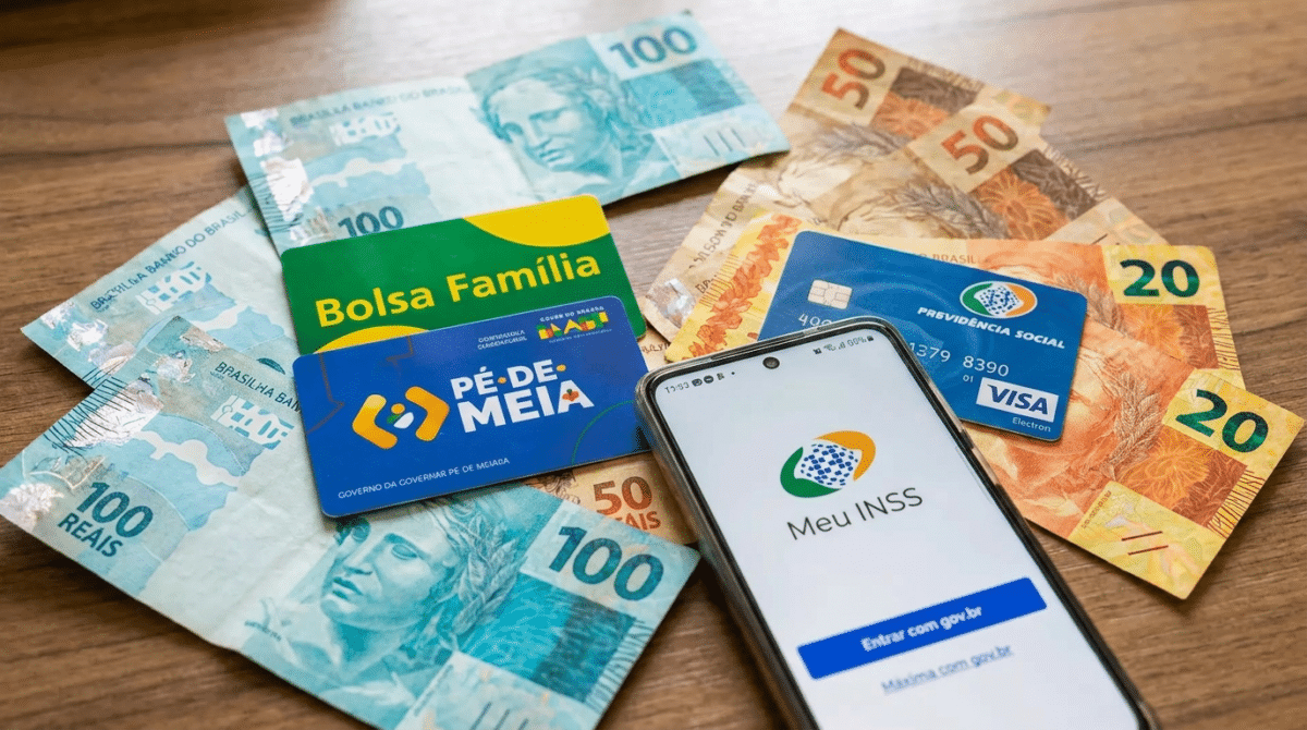 Cédulas de reais, cartões Bolsa Família e Pé-de-Meia com smartphone exibindo app Meu INSS sobre mesa de madeira.